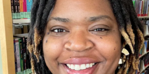 Damera Blincoe. A person with dreadlocks is standing in front of a bookshelf filled with various books. The individual is wearing a black top and has a necklace visible around their neck. The background features a library setting, with colorful book spines clearly visible on the shelves.