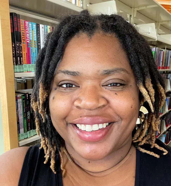 Damera Blincoe.
A person with dreadlocks is standing in front of a bookshelf filled with various books. The individual is wearing a black top and has a necklace visible around their neck. The background features a library setting, with colorful book spines clearly visible on the shelves.