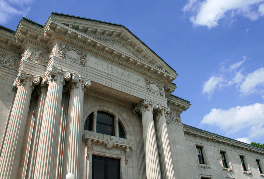 The image shows the exterior of a grand library building. The structure features classical architectural elements, including tall, fluted columns with ornate capitals. Above the entrance, the word "LIBRARY" is engraved prominently. The building has a triangular pediment with decorative details, and the facade is made of light-colored stone. The sky above is blue with scattered clouds, enhancing the overall aesthetic of the scene.