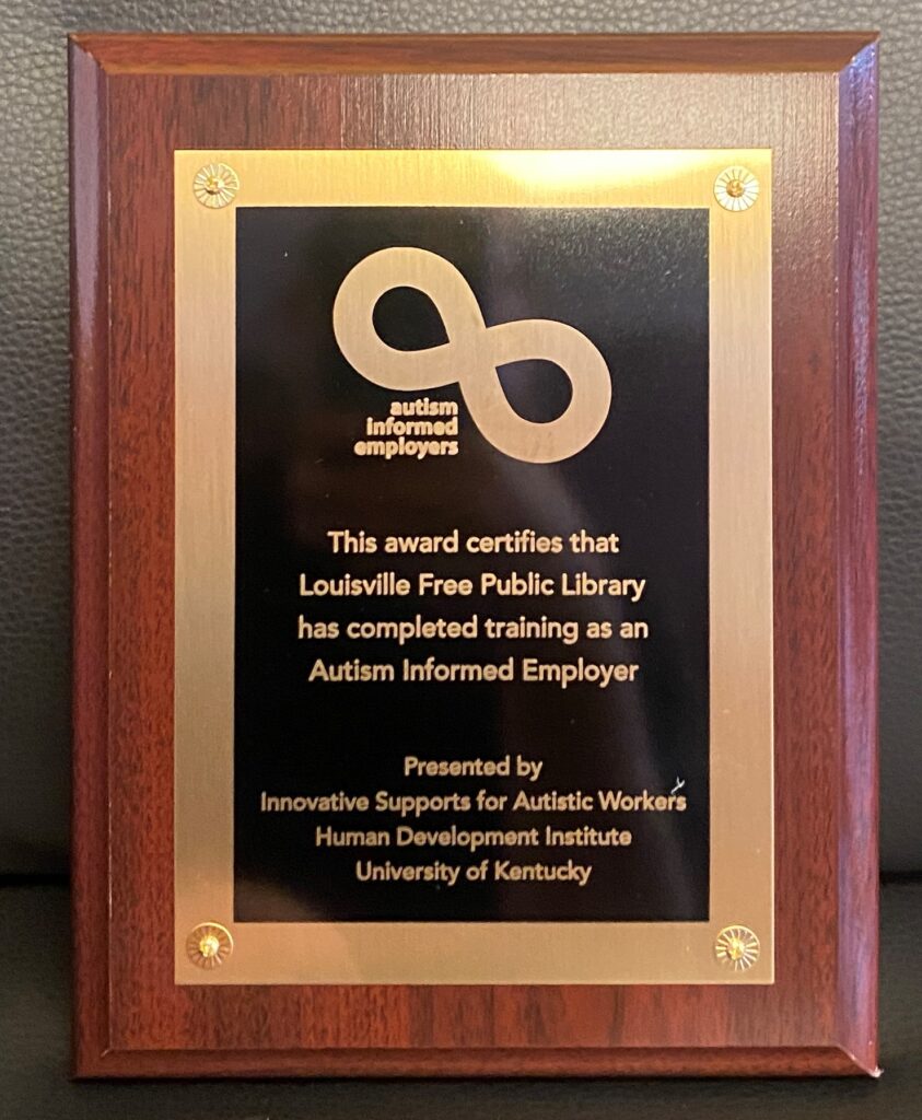 A black and gold plaque on wood.
 'လ autism Informed employers 
This award certifies that Louisville Free Public Library has completed training as an Autism Informed Employer. 
Presented by Innovative Supports for Autistic Workers Human Development Institute University of Kentucky'