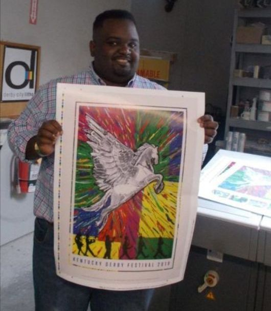 A man with dark skin and close cropped dark hair and beard smiles while holding a large print of a white pegasus flying with colorful background. 
