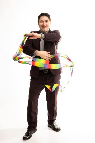 A man with light skin and short, dark hair wearing a dark brown suit while smiling at the camera and waving a rainbow colored ribbon in front of him.