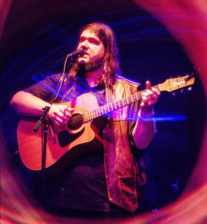 Photo of a man with light skin, long straight brown hair and beard holding a light brown acoustic guitar and singing into a microphone. 