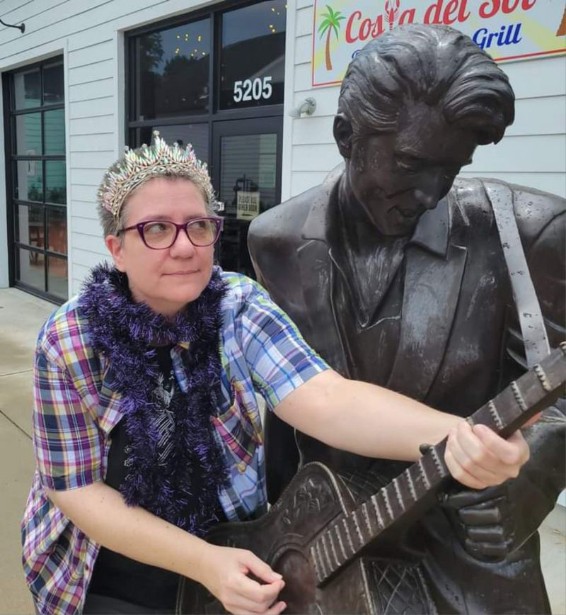 Person with light skin and glasses wearing a blue plaid shirt, a tiara, and a blue boa playfully pretending to play a guitar held by an Elvis statue.